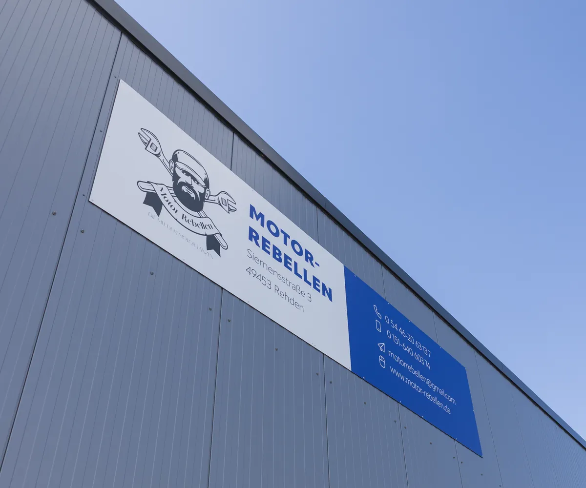 Aussenansicht der Werkstatt, Firmenschild ist sichtbar vor blauem Himmel